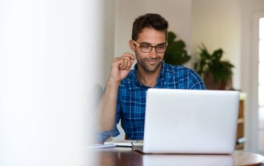 Genç adam yemek masasında oturuyor ve uzaktan bilgisayarla çalışırken gülümsüyor.