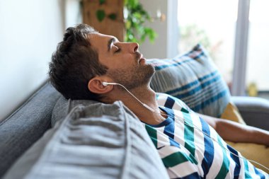 Genç adam oturma odasındaki kanepesinde gözleri kapalı ve kulaklıkla müzik dinliyor.
