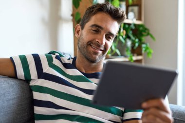 Gülümseyen genç adam oturma odasındaki kanepesinde oturuyor ve dijital tablet kullanıyor.