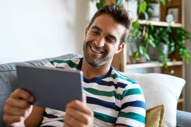 Evdeki kanepede otururken dijital tablet kullanan gülen genç adam.