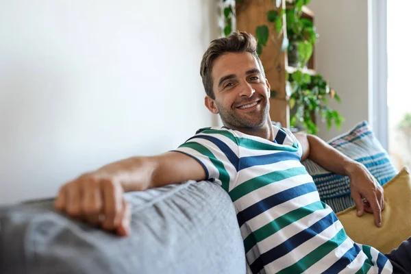 Portrait of a young man smiling while sitting alone on a sofa in his living room at home in the afternoon