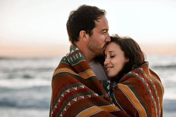 夕暮れ時に毛布に包まれたビーチで一緒に立っている間 若い男は妻にキスをしました — ストック写真