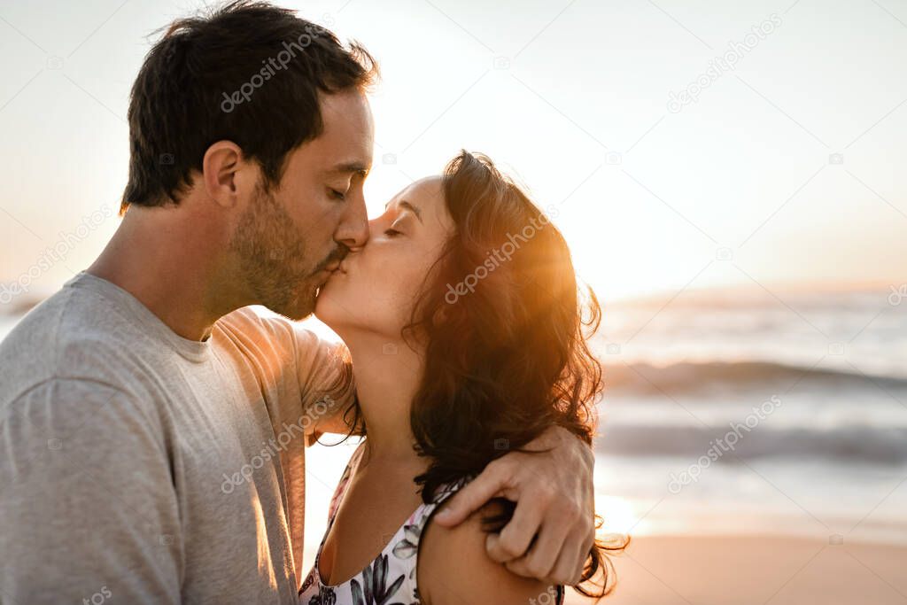 Loving young couple standing in each other's arms and kissing on a sandy beach at sunset