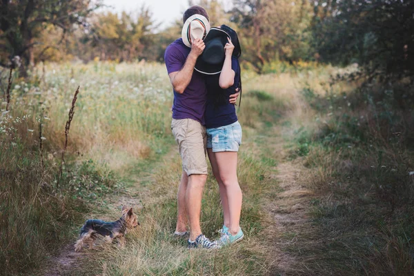 Молода пара з маленьким собакою йоркширським тер'єром одягнена в футболку і капелюх, що обіймає і розважається в дорозі — стокове фото