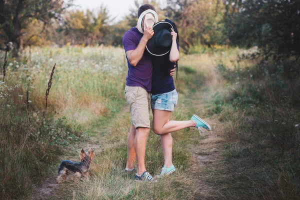 Молода пара з маленьким собакою йоркширським тер'єром одягнена в футболку і капелюх, що обіймає і розважається в дорозі — стокове фото
