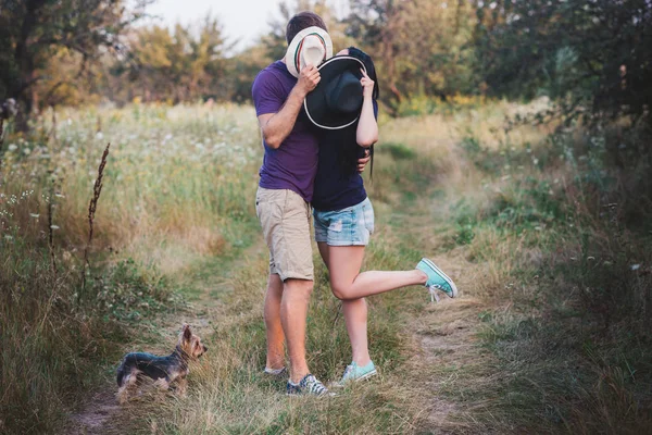 Молодая пара с маленьким собачьим йоркширским терьером, одетая в футболку и шляпу, обнимается и веселится на дороге — стоковое фото