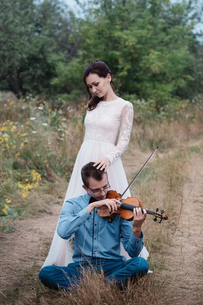 Hegedűművész és egy nő fehér ruhában, fiatal ember játszik a hegedű a háttérben természet, — Stock Fotó