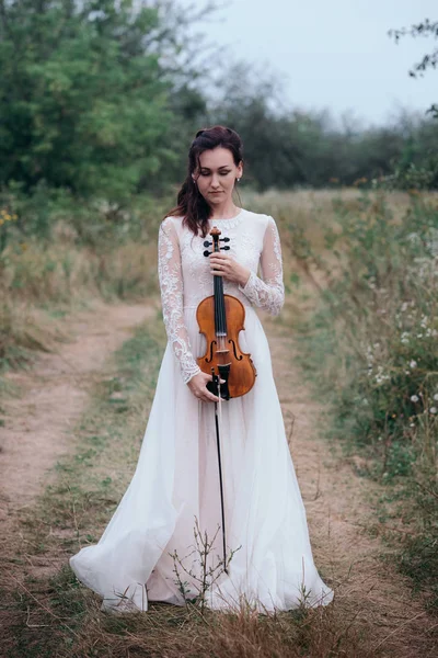 Joven chica hermosa en un vestido blanco bajo el árbol sosteniendo un violín en las manos . — Foto de Stock