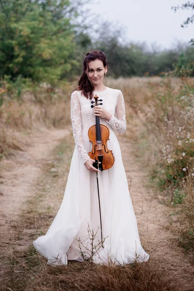 Fiatal gyönyörű lány hegedű tartja kezében fa alatt fehér ruhát. — Stock Fotó