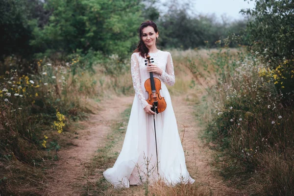 Bir keman elinde tutan ağaç altında beyaz elbiseli genç güzel kız.