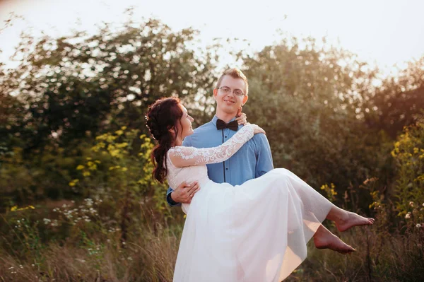 Γαμπρός νύφη μεταφέρει στην αγκαλιά του φύση με το φως του ηλιοβασιλέματος — Φωτογραφία Αρχείου