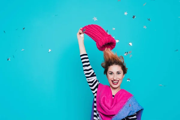 Stylish young woman removes a knitted pink hat from her head in the studio on a blue background. Winter goodbye. Spring comes. Lifestyle, youth, fun