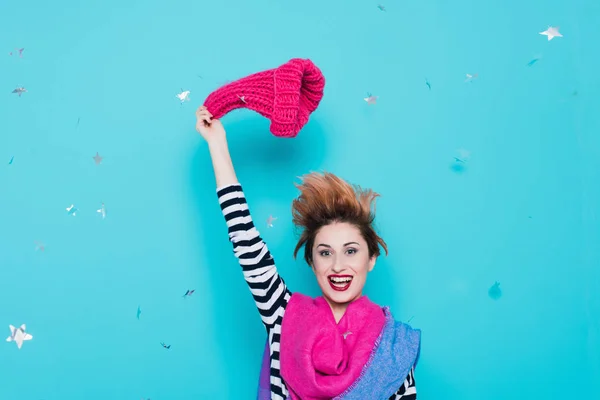 Stylish young woman removes a knitted pink hat from her head in the studio on a blue background. Winter goodbye. Spring comes. Lifestyle, youth, fun