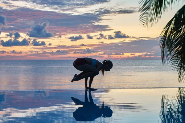 Young woman practices yoga on sea coastline during colorful sunset, travel concept