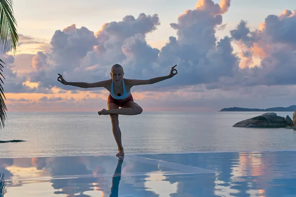 Young woman practices yoga on sea coastline during colorful sunset, travel concept