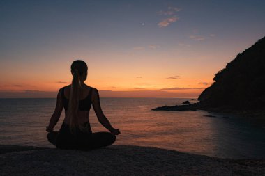Günbatımı arka planında deniz kıyısında meditasyon yapan genç bir kadının dikiz görüntüsü 