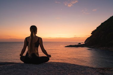 Günbatımı arka planında deniz kıyısında meditasyon yapan genç bir kadının dikiz görüntüsü 