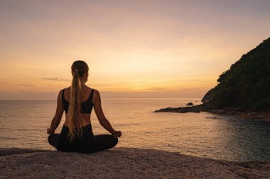 Günbatımı arka planında deniz kıyısında meditasyon yapan genç bir kadının dikiz görüntüsü 