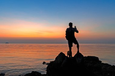 Sahildeki erkek fotoğrafçının silueti renkli günbatımını çekiyor.
