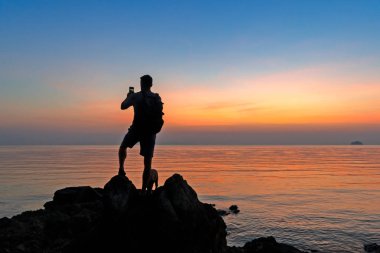 Sahildeki erkek fotoğrafçının silueti renkli günbatımını çekiyor.