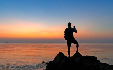 Sahildeki erkek fotoğrafçının silueti renkli günbatımını çekiyor.