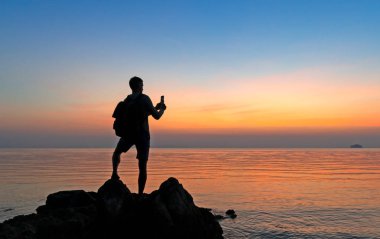 Sahildeki erkek fotoğrafçının silueti renkli günbatımını çekiyor.
