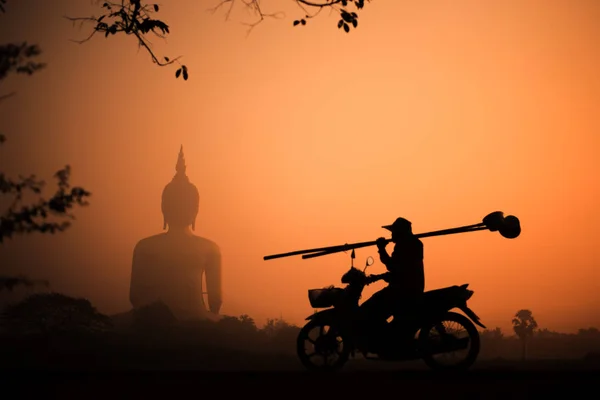 Silhouette big Buddha with the golden sky on the sunset and silhouette of man riding a motorcycle holding bamboo stick afterwork.