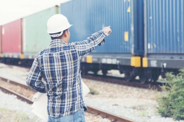 Akıllı mühendis elinde beyaz başlık tutuyor ve fabrika merkezindeki rafineri fabrikasına bakıyor. Asyalı adam demiryolu inşaat mühendisliği konseptini el ele veriyor.