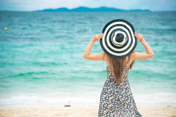 Summer time tan woman vacation on the beach. Cheerful woman wear summer dress and straw hats sitting on the beach look at sea. Time to relax in summer lifestyle outdoor shot on tropical island beach.