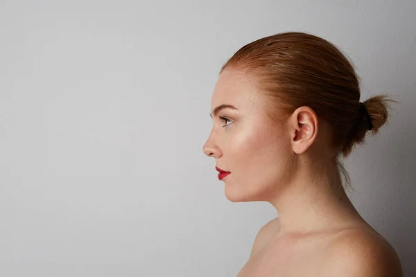 Side portret van mooie jonge vrouw met naakte schouders en rode lippen kijken naar de camera en glimlachen, poseren op de grijze muur. geïsoleerd. — Stockfoto