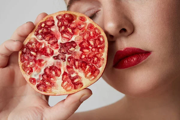 Close-up portret van mooie jonge vrouw met rode lippen en gesloten ogen met verse granaatappel en glimlachend, poserend op de grijze muur. geïsoleerd. — Stockfoto