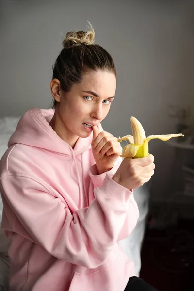 Junge Frau sitzt im Bett, isst Banane und ruht sich aus. Vertikales Bild. — Stockfoto