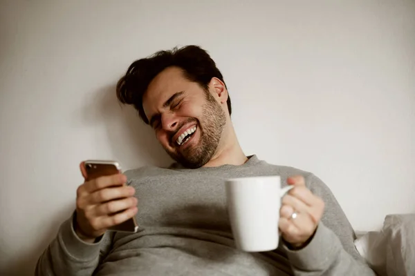 Bearded hipster man lays down laughing relaxing on his bed talking to someone. Hipster, conversing. Space for text.