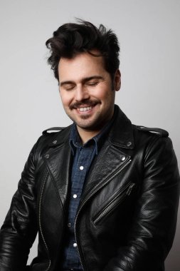 Motosiklet ceketi giyen komik sakallı adam tuhaf bir şekilde gülümsüyor ve kameraya kapalı gözlerle bakıyor. Dikey çekim.