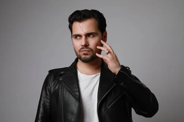 Foto de la cabeza de un joven hipster moderno y elegante tocándose la cara con una chaqueta de cuero posando sobre un fondo blanco . — Foto de Stock