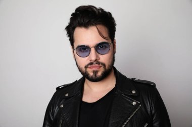 Deri elbiseli, parlak beyaz tişörtlü, şık gözlüklü, beyaz duvarda poz veren sakallı bir adam. İzole edilmiş.