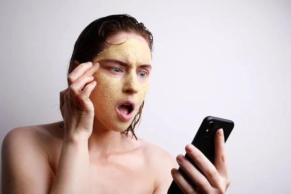 Detailní záběr portrét mladé bělošky s překvapivou tváří nanášení obličejové hliněné masky pomocí jejího smartphonu. — Stock fotografie