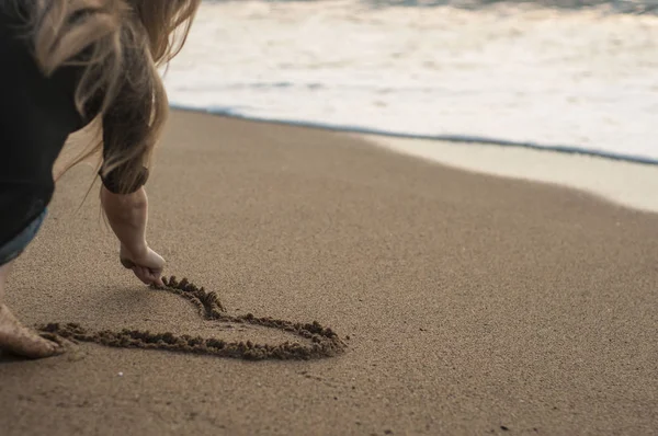 Mladá dívka nakreslí srdce v písku, sedí na pláži — Stock fotografie