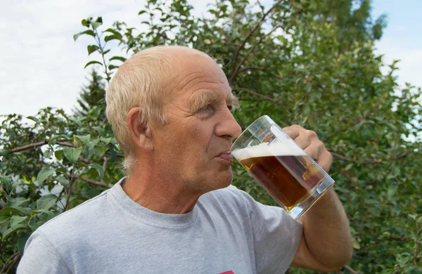 An elderly man with delight and pleasure is drinking cold beer from a mug on a background of trees in your garden