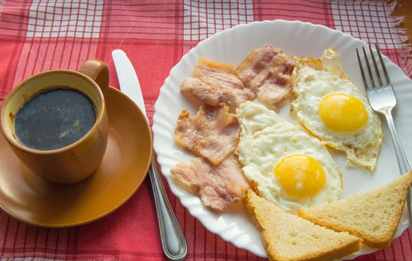 Вкусный завтрак - чашка кофе, тарелка жареных яиц, бекон и тост, рядом со столовыми приборами на красной клетчатой салфетке — стоковое фото