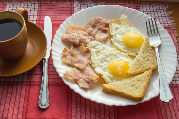 Finom reggeli - egy csésze kávé, egy tányér sült tojás, szalonna és pirítós, mellett az evőeszközök piros kockás szalvéta — Stock Fotó