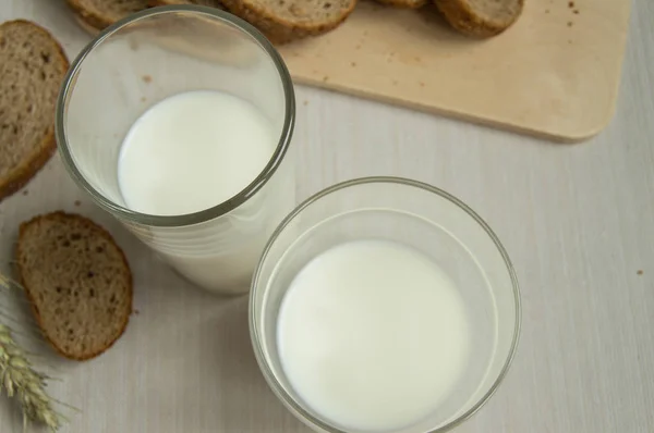 Dois copos de leite ao lado de uma tábua de madeira e pão na mesa, vista superior — Fotografia de Stock