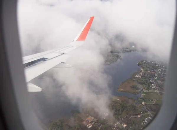 Uma visão panorâmica da asa nas nuvens e do solo com casas através da janela da aeronave durante o voo — Fotografia de Stock