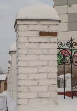 Beyaz alçılı tuğla sütunlar arka arkaya metal parmaklıklarla çit oluşturuyorlar. Sütunların üzerinde büyük kar yığınları, kentsel kış arkaplanı var.