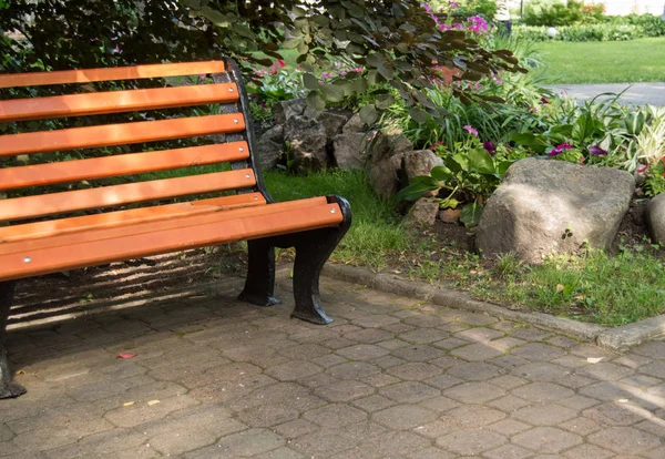 Часть деревянной скамейки возле клумбы с цветами и камнями в солнечный летний день в городском парке — стоковое фото