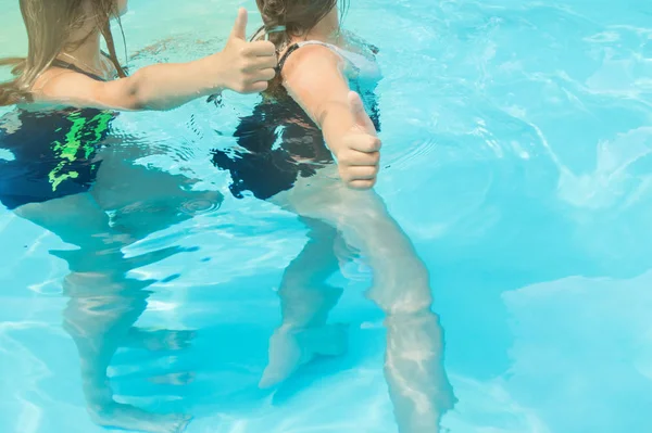 Пальцы вверх две девочки-подростки с длинными волосами в купальниках в бассейне с голубой игристой водой на открытом воздухе, солнечный свет, яркое лето — стоковое фото