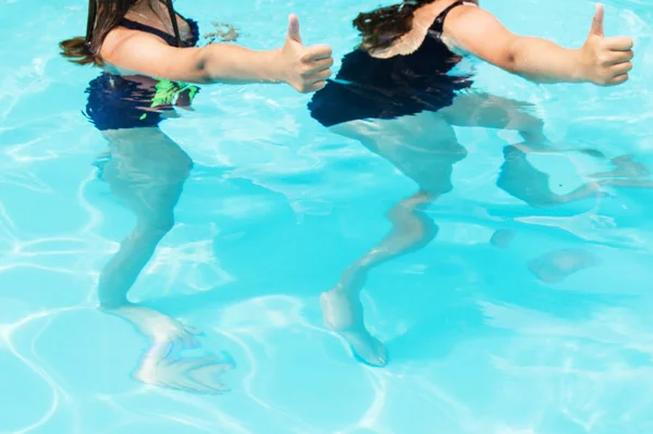 Пальцы вверх две девочки-подростки с длинными волосами в купальниках в бассейне с голубой игристой водой на открытом воздухе, солнечный свет, яркое лето — стоковое фото