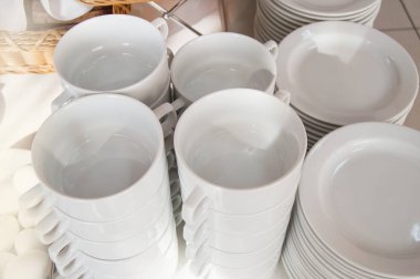 Beyaz seramik çay fincanları ve tabaklar bir masanın üzerinde beyaz bir masa örtüsü ve konukların kahvaltısı için hazırlanan otel restoranındaki temiz tabaklar yakın plan.
