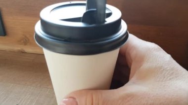 A woman's hand takes a paper Cup of coffee or tea in a cafe on a wooden table with a view of the street and random passers-by on a blurred background. Hand turns the glass