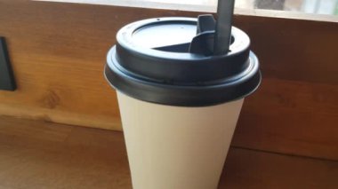 There is a white paper Cup of coffee on a wooden table in the cafe, and through the glass you can see the street with a blurred focus.